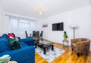A modern living room with a blue sofa, a glass coffee table, a dining table with chairs, a flat-screen TV, and a floor lamp. - Spacious 3BR S/E London Flat in Penge with Parking sleeps 6 (London)