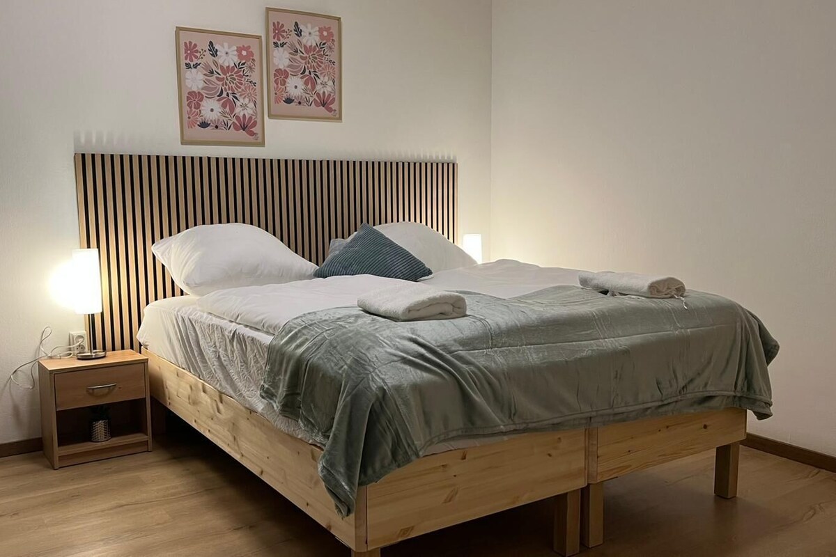 A neatly made bed with a wooden headboard, a bedside table with a lamp, and two floral wall art pieces.