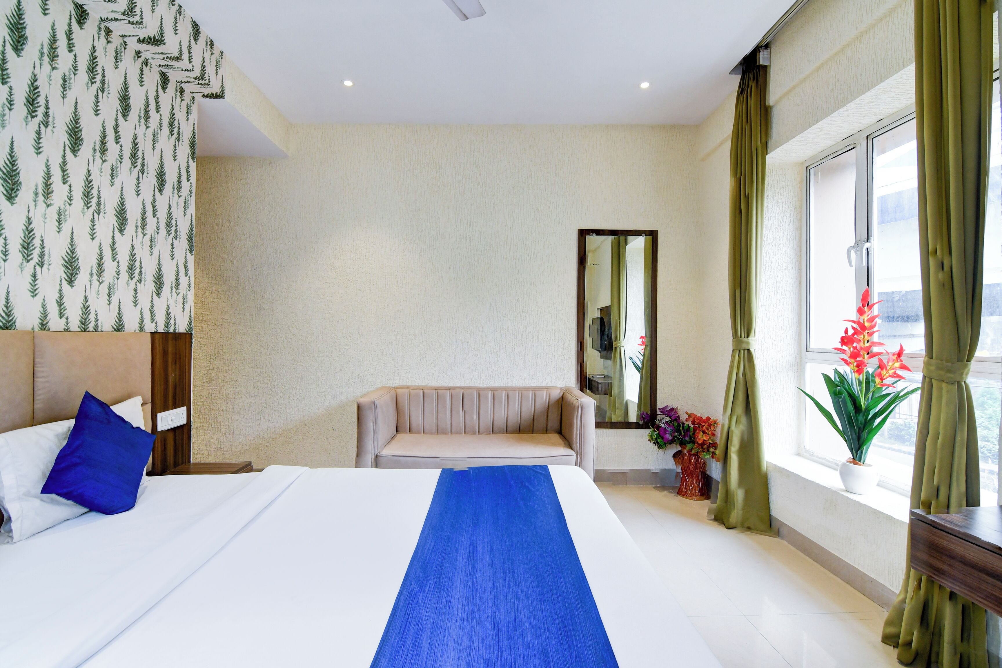Una habitación de hotel con cama, sofá, espejo, ventana con cortinas y vistas al exterior.