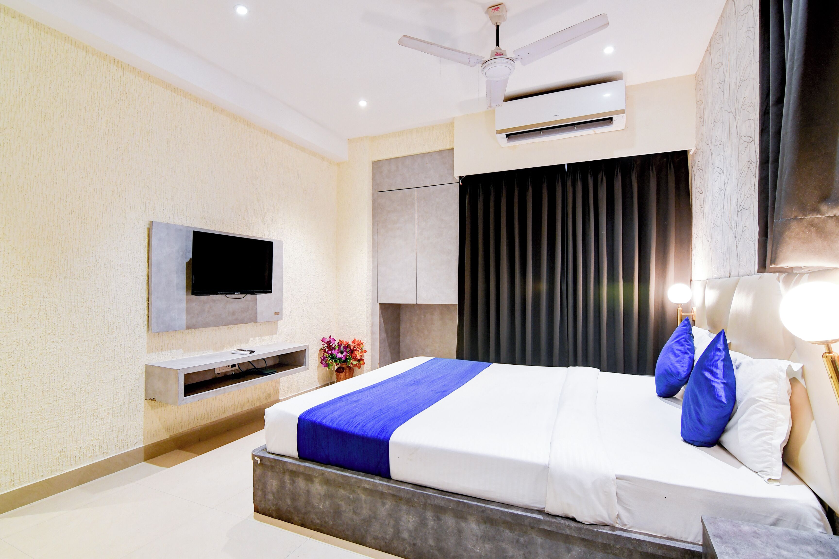 A modern hotel room with a large bed, a flat-screen TV, a ceiling fan, and a built-in air conditioning unit.