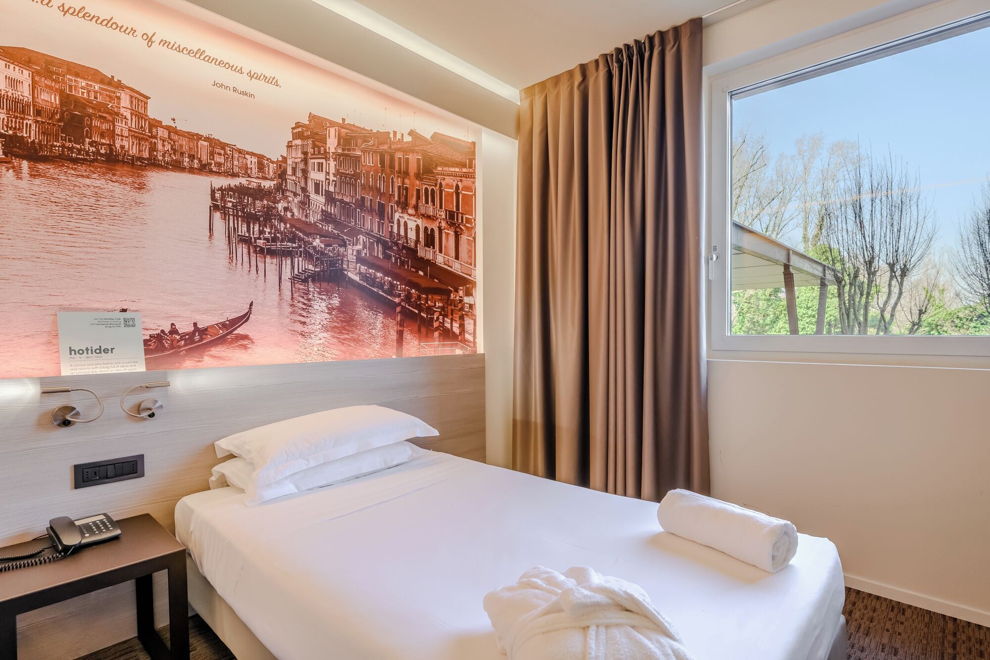 A hotel room with a bed, a telephone, a poster of a cityscape, and a window with curtains.