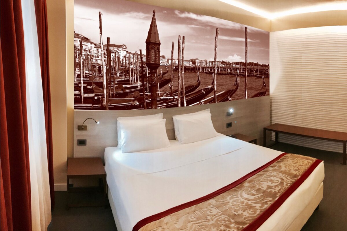 A hotel room with a large bed, a bench, a wall-mounted TV, and a door leading to a bathroom.