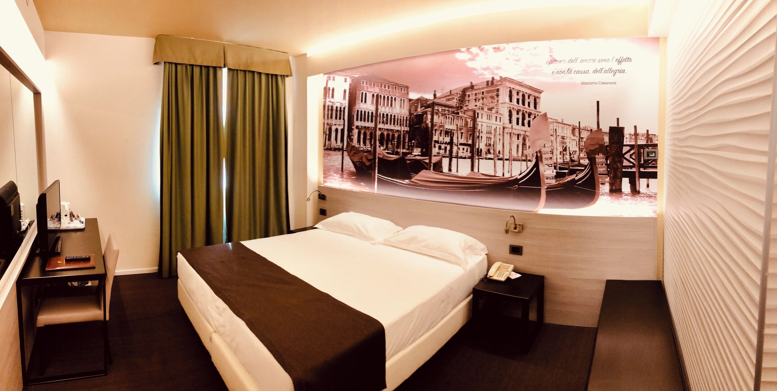 A hotel room with a bed, a desk with a chair, a nightstand with a phone, a window with curtains, and a wall with a framed picture.