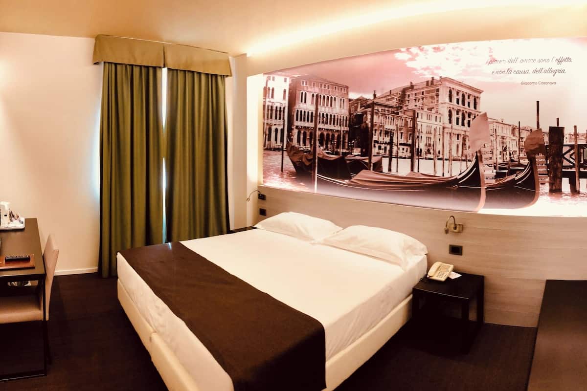 A hotel room with a bed, a desk with a chair, a nightstand with a phone, a window with curtains, and a wall with a framed picture.