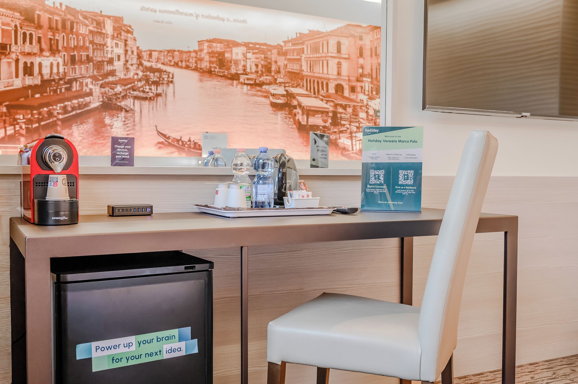 Een hotel receptiebalie met een koffieapparaat, waterflessen, een prullenbak en een stoel.