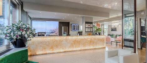 Eine moderne Hotelhalle mit einer Marmor-Rezeption, einem grünen Sofa, einem weißen Sessel und einem großen Fenster mit Blick auf die Stadt.