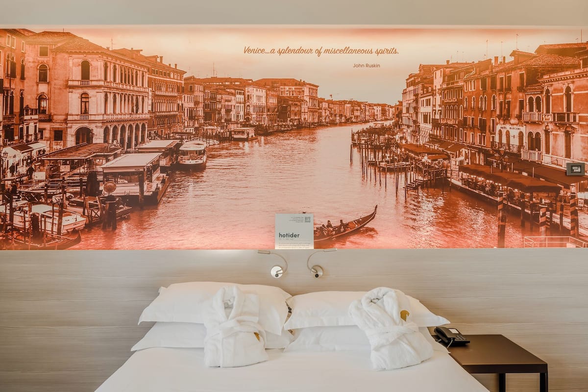 A hotel room with a large poster of Venice's Grand Canal, a bed with white linens, two bedside tables, and a single bedside lamp.