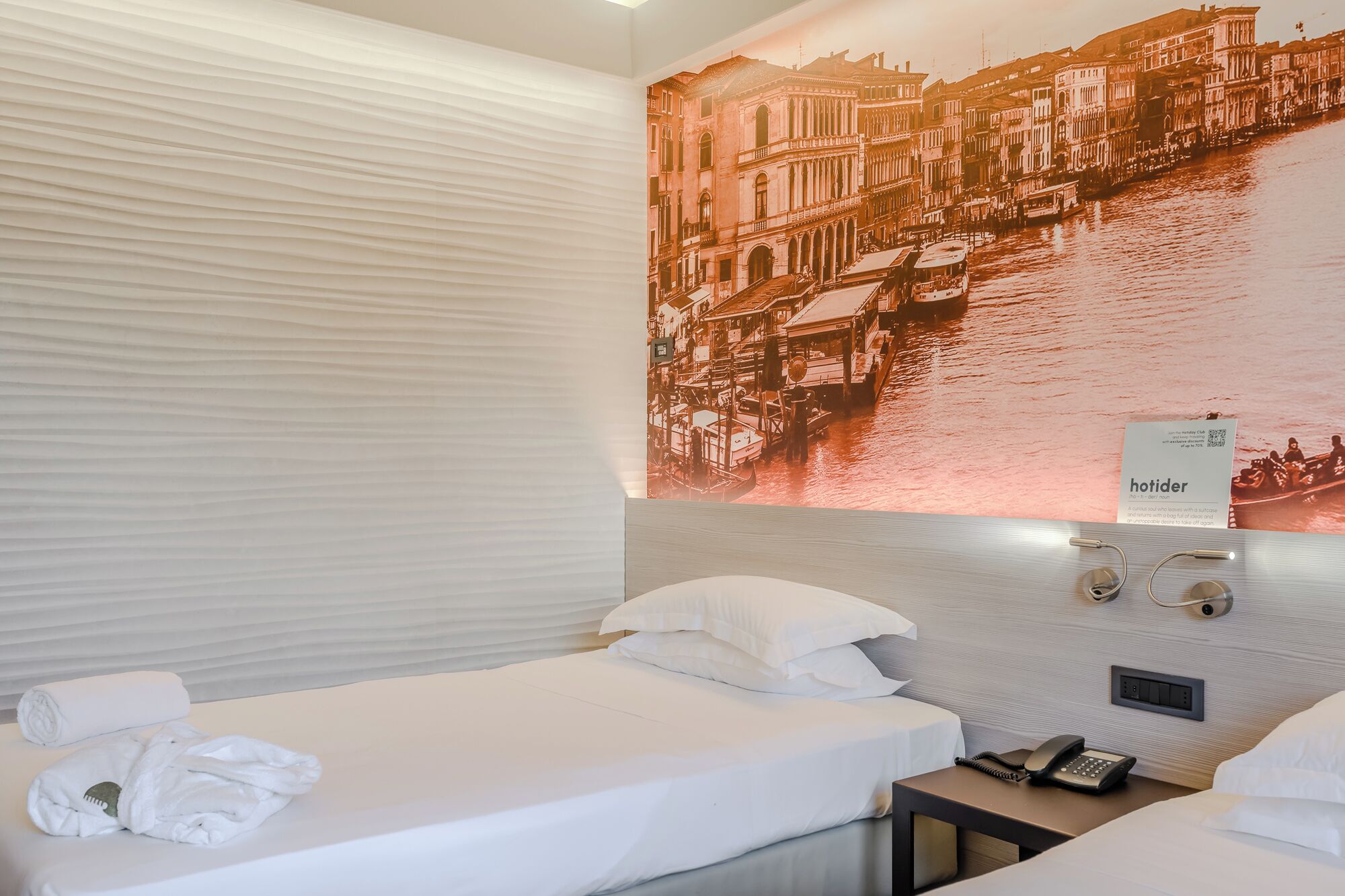 A hotel room with two beds, a large wall mural of a cityscape, a sign reading "hotider," and a telephone on the nightstand.