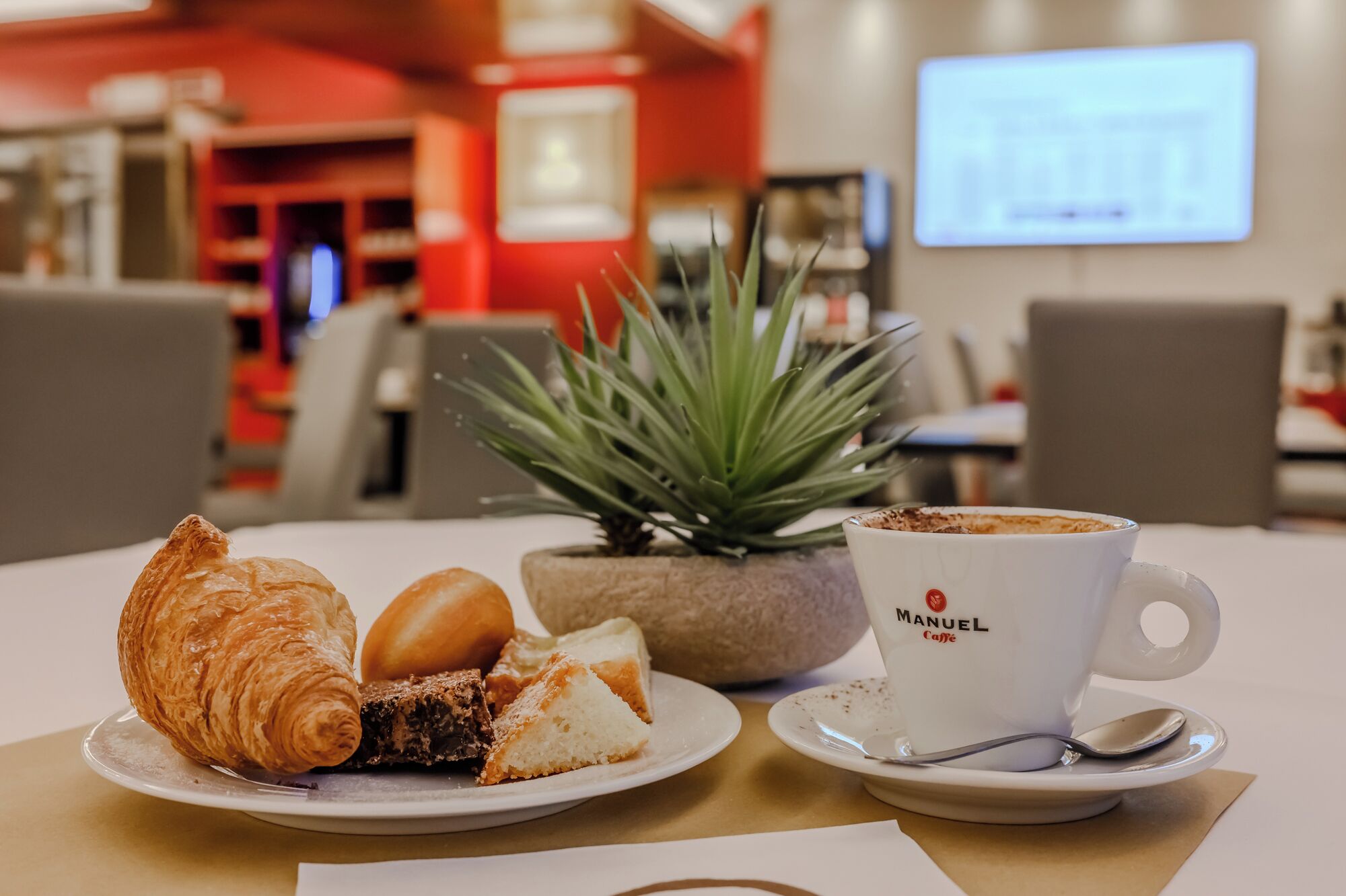 A table with a croissant, a slice of cake, and a cup of coffee labeled 'Manuel Caffe'. There is a potted plant and a screen displaying a graph in the background.