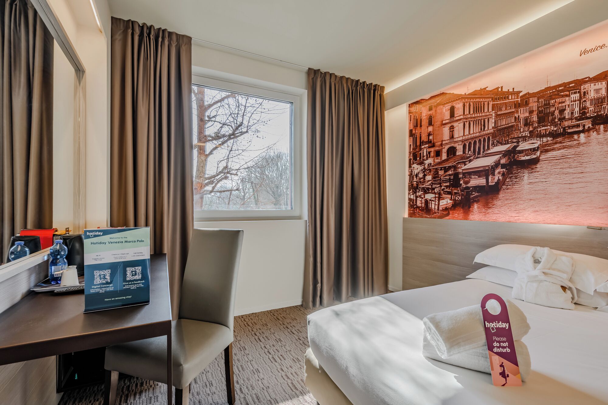 Une chambre d’hôtel avec un lit, un bureau et une chaise, une fenêtre avec des rideaux et un mur orné d’une affiche représentant un paysage urbain.