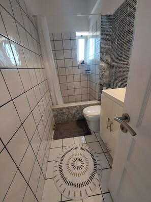Casa de banho com paredes em azulejo branco, zona de duche, sanita e lavatório.