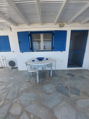 Uma área de refeições ao ar livre coberta, com piso de pedra, uma mesa redonda e persianas azuis nas janelas.