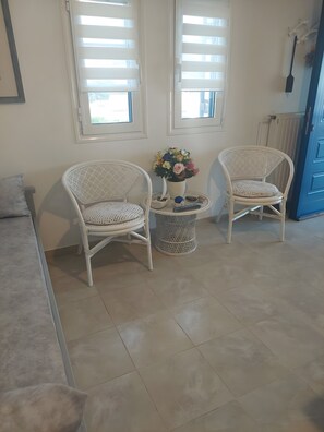 A room with two wicker chairs, a small round table with a vase of flowers, and a blue door in the background. - Xanthi House Sikinos (Σίκινος)