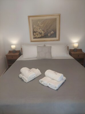 Cama bem arrumada com lençóis brancos, duas luminárias de cabeceira, um quadro com flores na parede e duas toalhas enroladas sobre a cama.