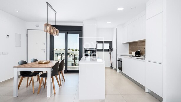 A modern kitchen with white cabinets, a wooden dining table, and a view of the city through large windows. - Golf Vista Penthouse, golf views, rooftop dining - by Oyoostays (Pilar de la Horadada)