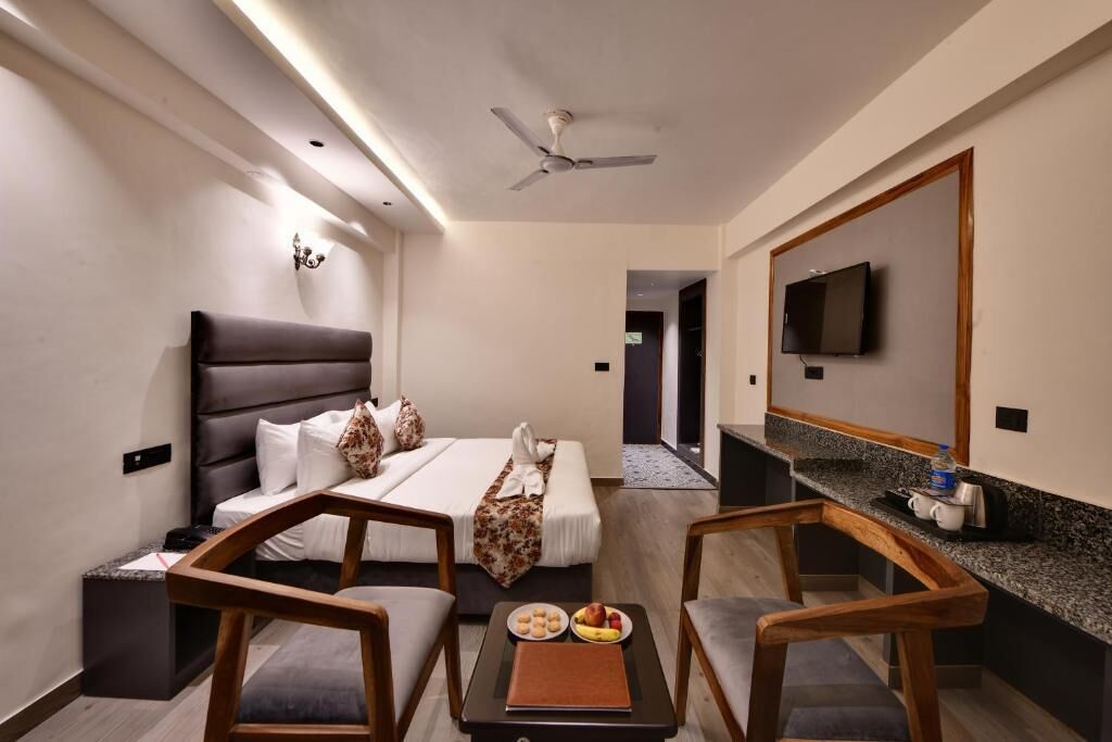 A hotel room with a bed, two chairs, a small table with a book and fruit, a TV, and a mirror.