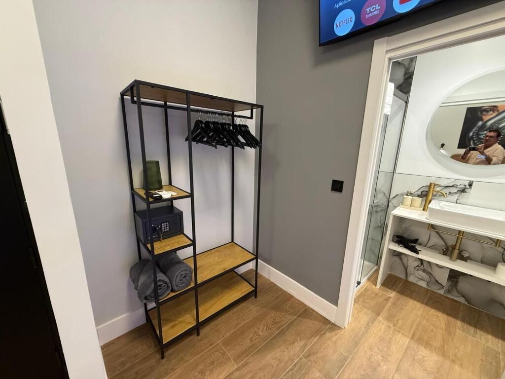 Une pièce avec un plancher en bois, une étagère en métal noir où sont rangés des vêtements et des serviettes, un téléviseur fixé au mur, et une salle de bain avec un miroir et un lavabo.