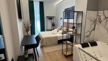 A compact hotel room with a bed, a small table, a chair, a desk, a bookshelf, and a bathtub.