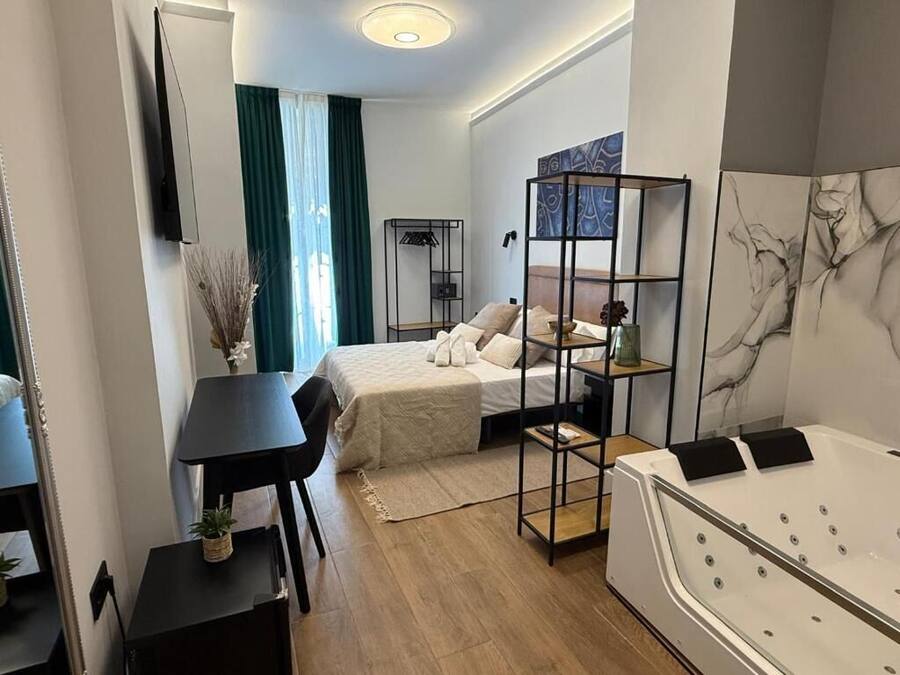 A compact hotel room with a bed, a small table, a chair, a desk, a bookshelf, and a bathtub.