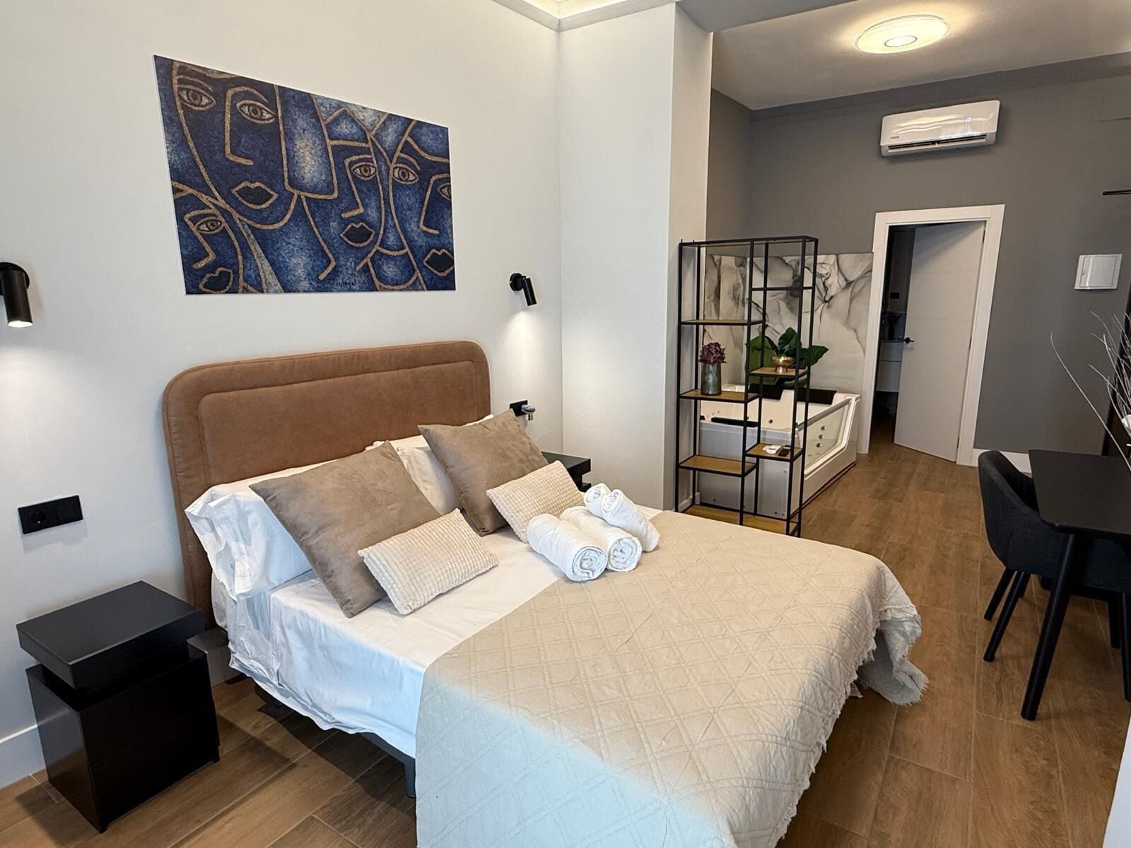 Un dormitorio con cama, mesita de noche, escritorio y silla. Hay un cuadro en la pared y un equipo de aire acondicionado en el techo.