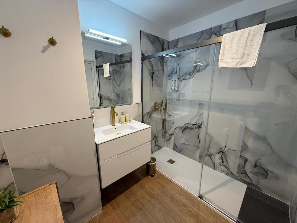 Un bany modern amb respatller de marbre, dutxa amb envàs de vidre i un moble de tocador blanc amb un faucet daurat.
