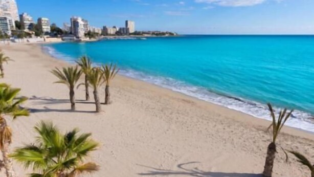 Una playa con agua turquesa cristalina, palmeras y edificios al fondo.