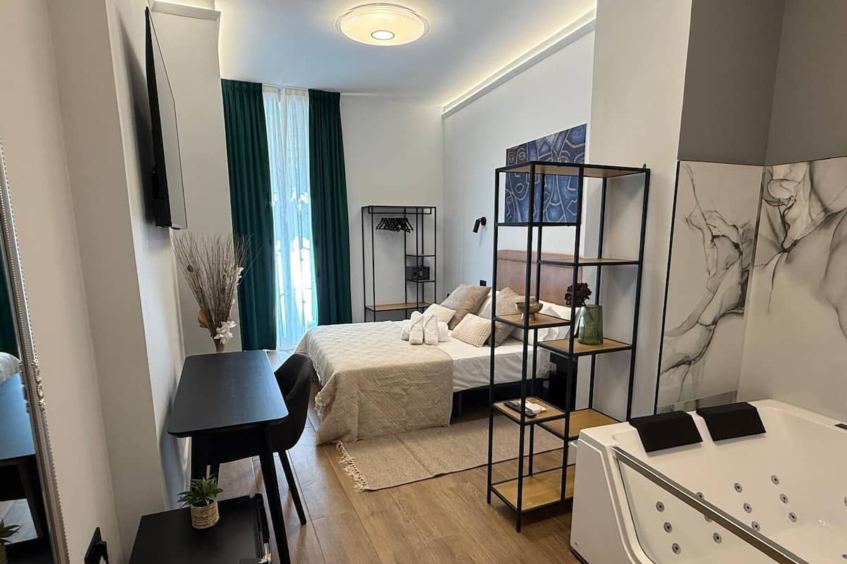 A modern hotel room with a bed, a desk, a chair, a small table, a sofa, a TV, and a bathroom with a bathtub.
