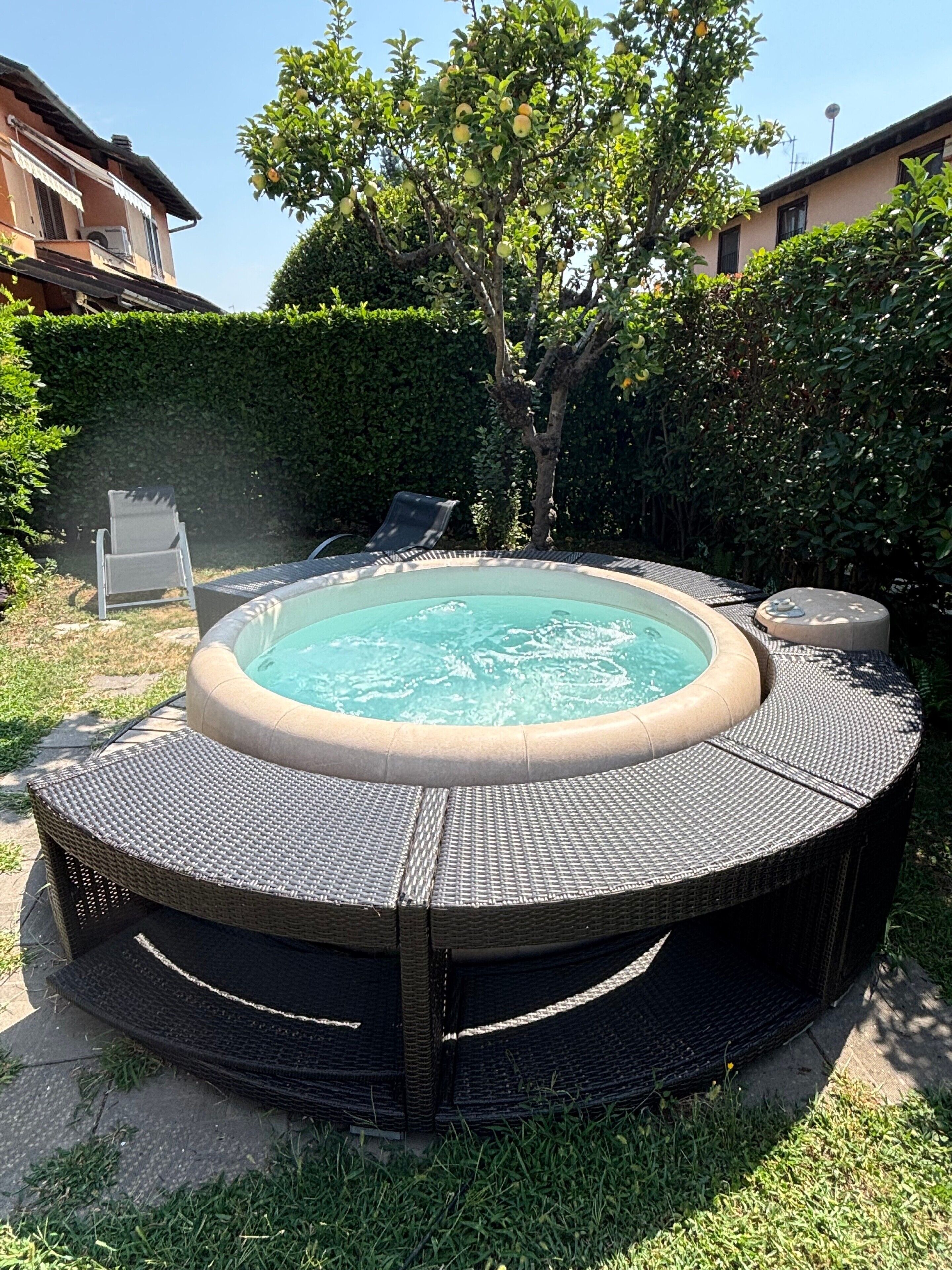 Ein Whirlpool, umgeben von einer Rattan-Terrasse, mit einem Baum und einer Hecke im Hintergrund.