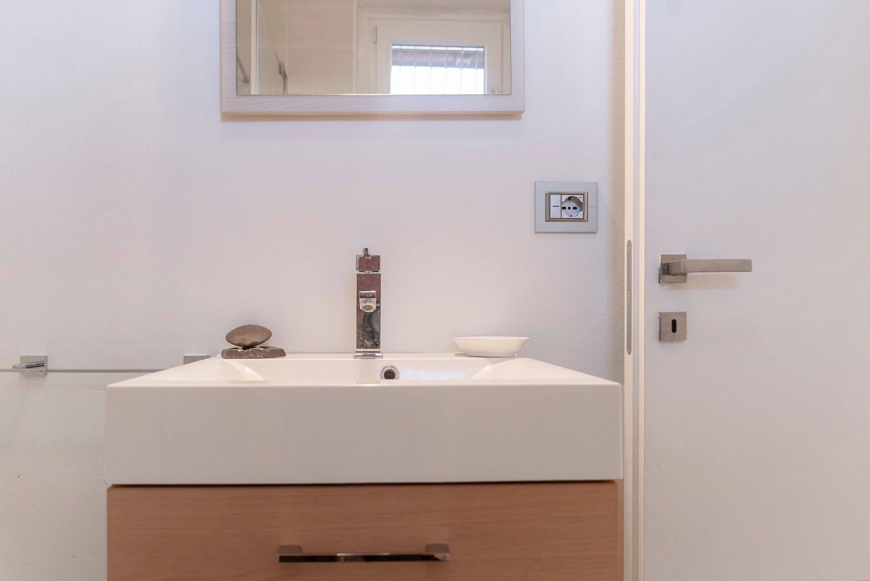 Une salle de bain avec un lavabo blanc, un miroir, un petit tableau encadré et une porte.