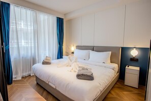 Une chambre d’hôtel moderne avec un grand lit, du linge de lit blanc, une table de chevet avec une lampe et une vue à travers des rideaux transparents.