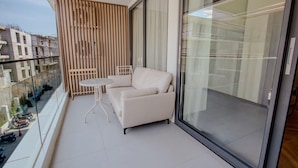 Ein Balkon mit einer weißen Couch, einem kleinen Tisch, einer Holzhocker-Abteilung und einer Glastür mit Blick auf die Stadt.
