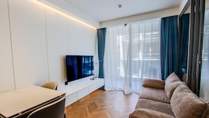 Ein modernes Wohnzimmer mit einem wandmontierten Flachbildfernseher, einer Couch, einem Esstisch mit Stühlen und einem großen Fenster mit transparenten Vorhängen.