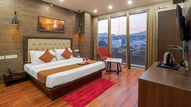 A modern hotel room with a large bed, a red rug, a desk with a kettle, and a city view through large windows. - Hotel Siya Ram by AKC Hotels (Haridwar)