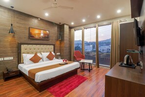 A modern hotel room with a large bed, a red rug, a desk with a kettle, and a city view through large windows. - Hotel Siya Ram by AKC Hotels (Haridwar)