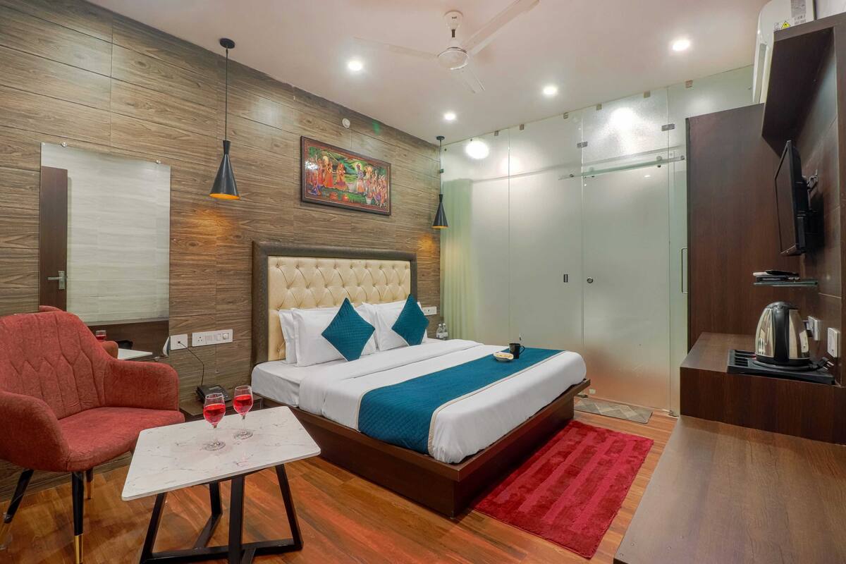 Una habitación de hotel con una cama, un sillón rojo, una mesita con dos copas de vino tinto, un tapete rojo y un televisor sobre un mueble de madera.