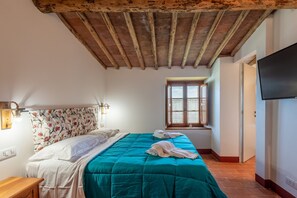 A bedroom with a wooden ceiling, a bed with a teal bedspread, a window with a view, and a flat-screen TV on the wall. - Room 'Castagno' – Agriturismo la Taverna di Celsa with Shared Pool (Sovicille)