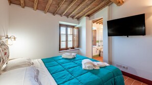 Un dormitorio con techo de madera, cama con colcha azul turquesa, televisor de pantalla plana en la pared y ventana con persianas.