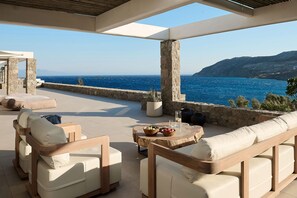 A sunlit outdoor seating area with stone pillars, a round wooden table with a bowl of fruit, and a view of the sea and hills. - Villa Socrate with pool and gym - My Rental Homes (Mykonos)
