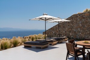 An outdoor seating area with a stone wall backdrop, a view of the sea, and a round table with chairs. - Villa Pegaso, pool and jacuzzi - My Rental Homes (Mykonos)