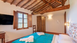 A bedroom with a wooden ceiling, a bed with a teal bedspread, a television on the wall, and a window with curtains. - Camera Castello – Agriturismo la Taverna di Celsa with Shared Pool (Sovicille)