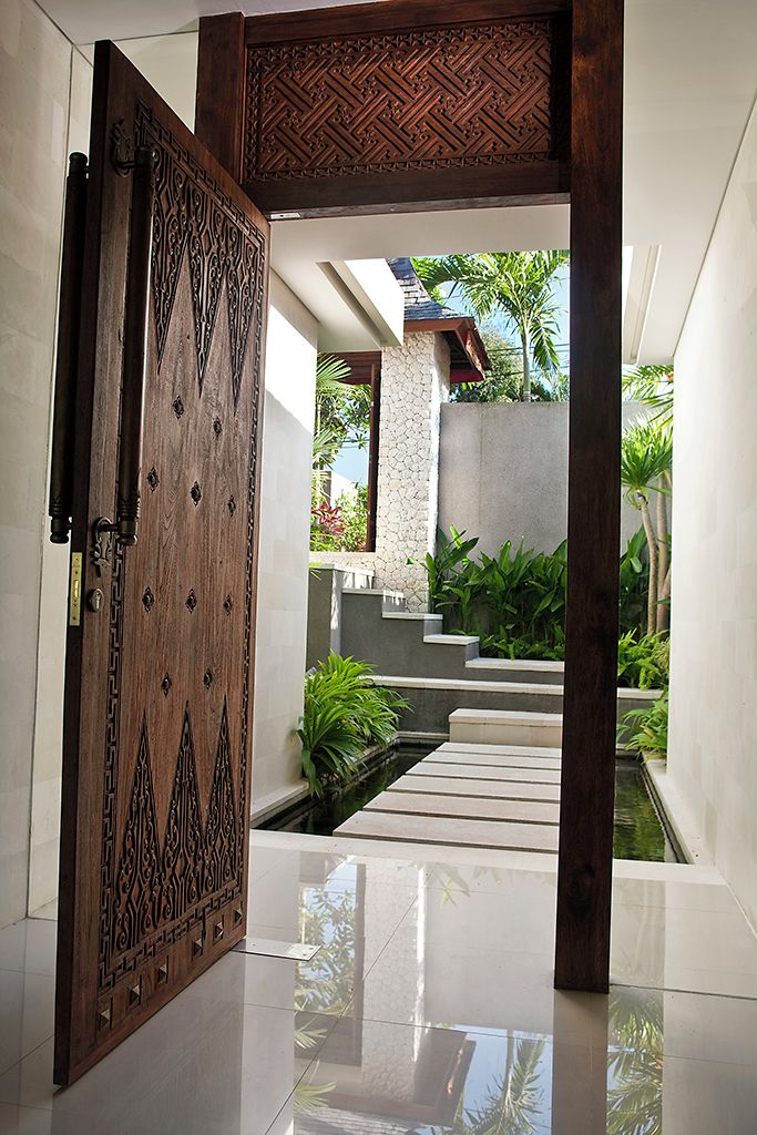 An open wooden door leading to a modern interior with a stone wall, a small water feature, and a view of a residential area with greenery.