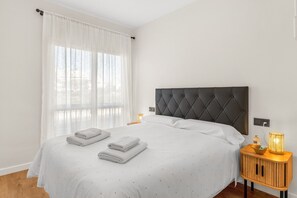A neatly made bed with a tufted headboard, white linens, and folded towels. There is a bedside table with a lamp and a small basket. - B&B Apartments. Modern and Luxurious Apartment Less Than 100 Meters from the Beach (La Cala de Mijas)