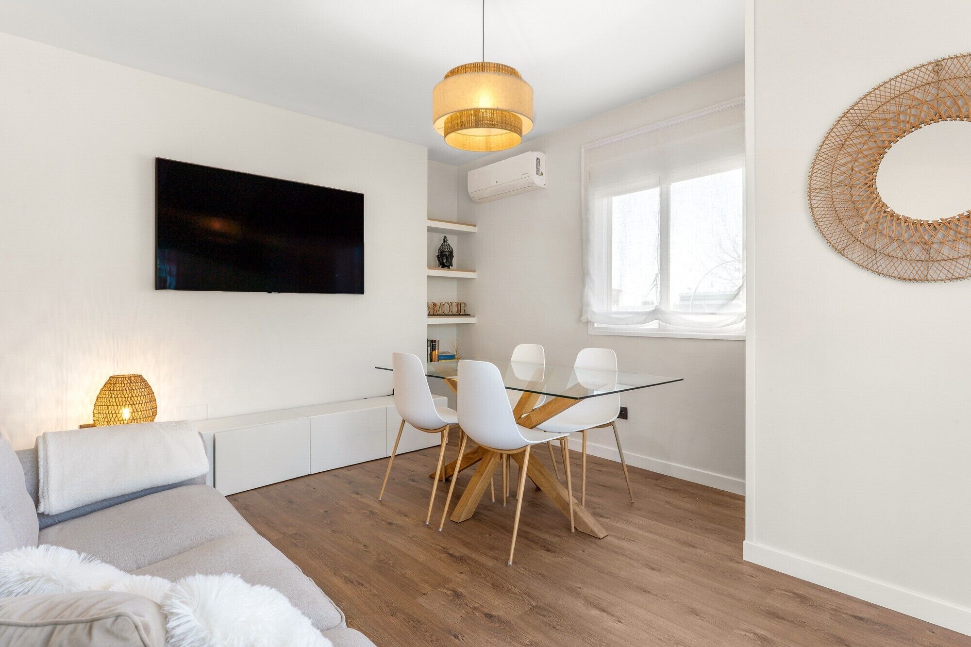 Una sala de estar moderna con televisor de pantalla plana, una mesa de comedor de vidrio con sillas blancas, una lámpara colgante y una ventana que deja entrar luz natural.