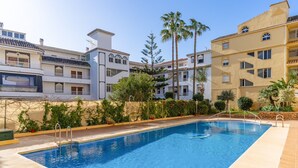 Una piscina amb aigua blava cristal·lina, envoltada per una paret de pedra baixa i al costat d'un edifici de diversos pisos amb balcons i un teulat llosat.