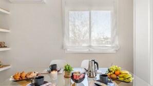 Zona de menjador moderna amb taula de vidre, cadires blanques, finestra amb cortines lleugeres i diverses coses per esmorzar.