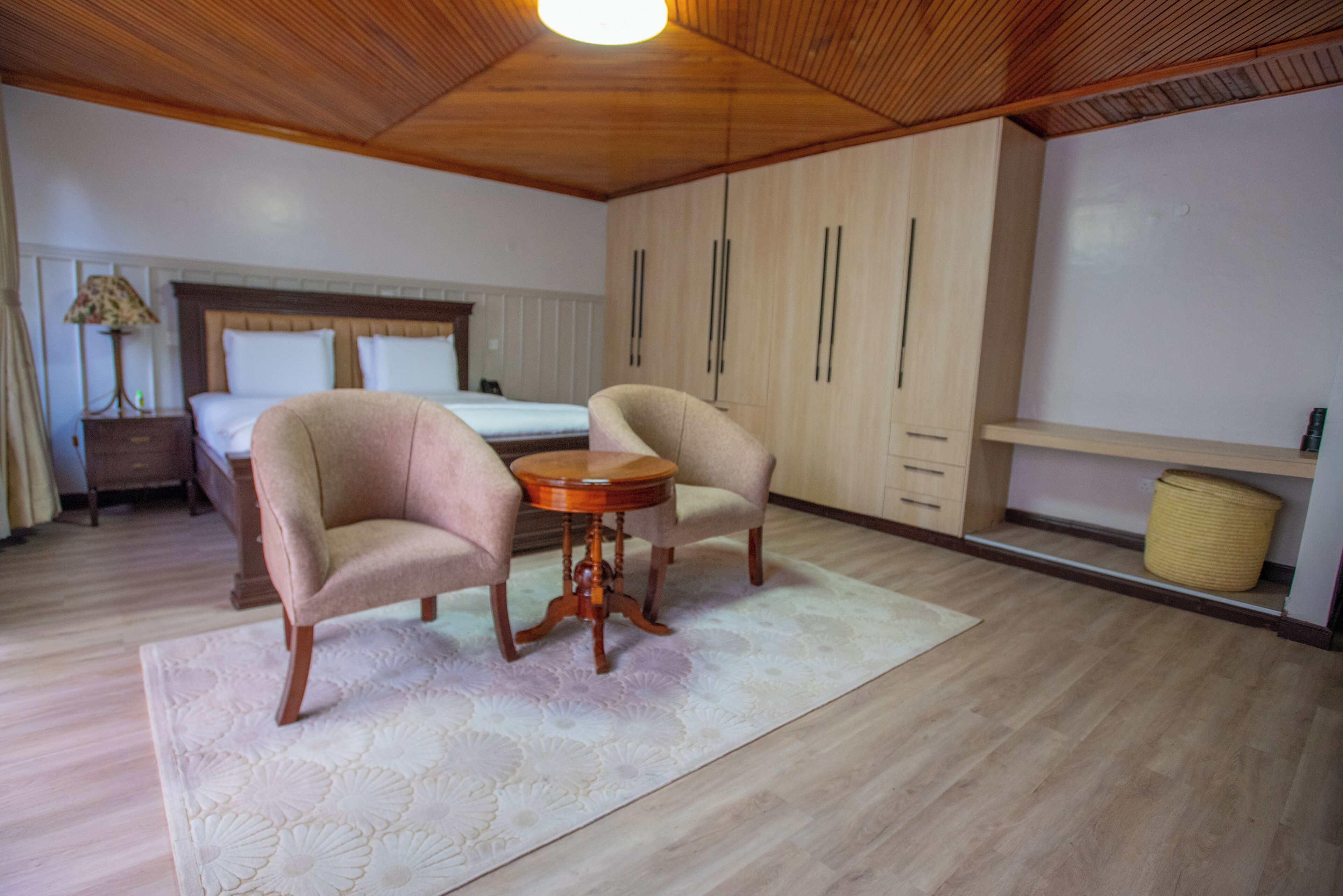 A bedroom with a bed, two armchairs, a round table, wooden wardrobe, and a nightstand with a lamp.