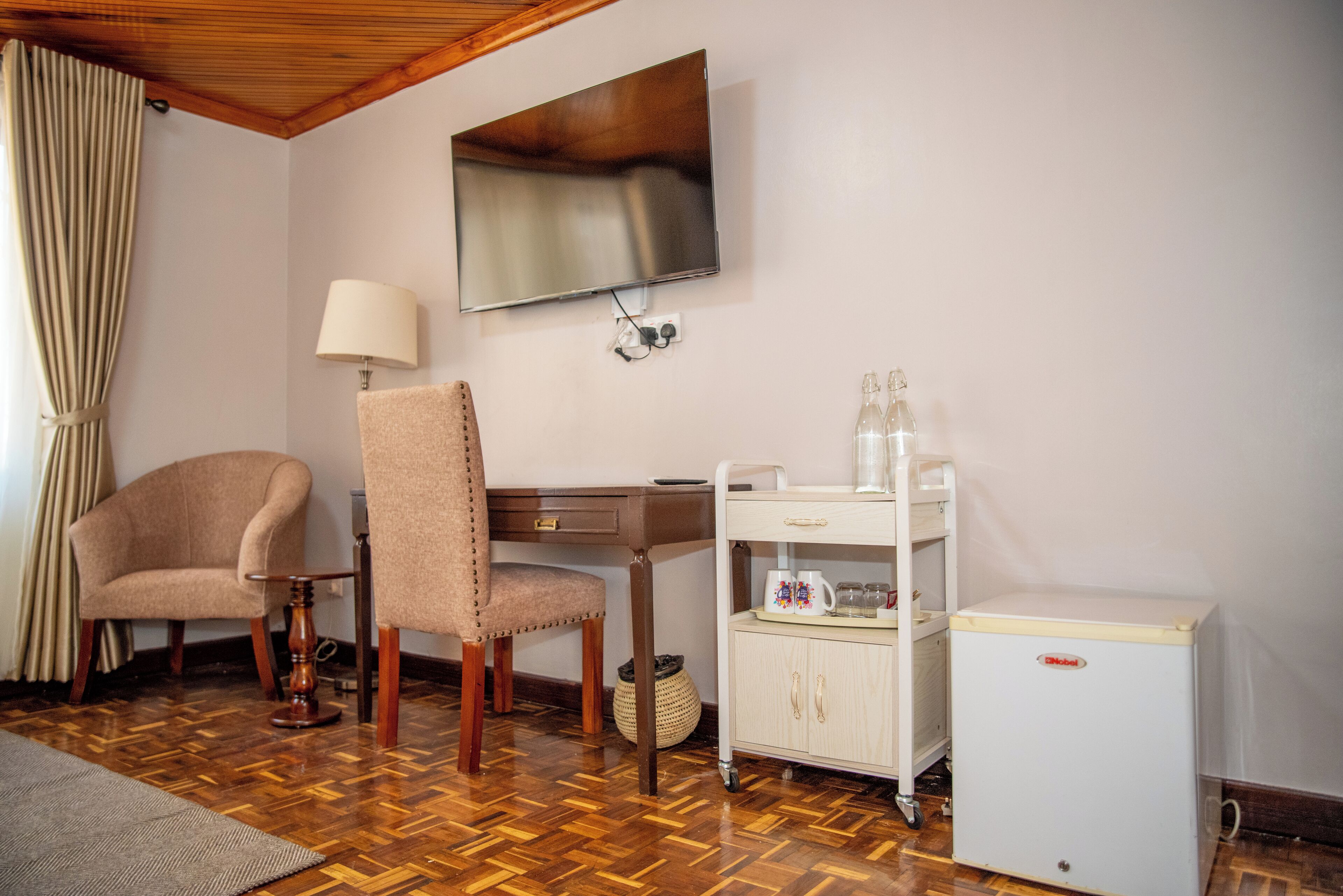 A room with a wooden floor, a TV mounted on the wall, a desk with a chair, a small refrigerator, and a cart with bottles.