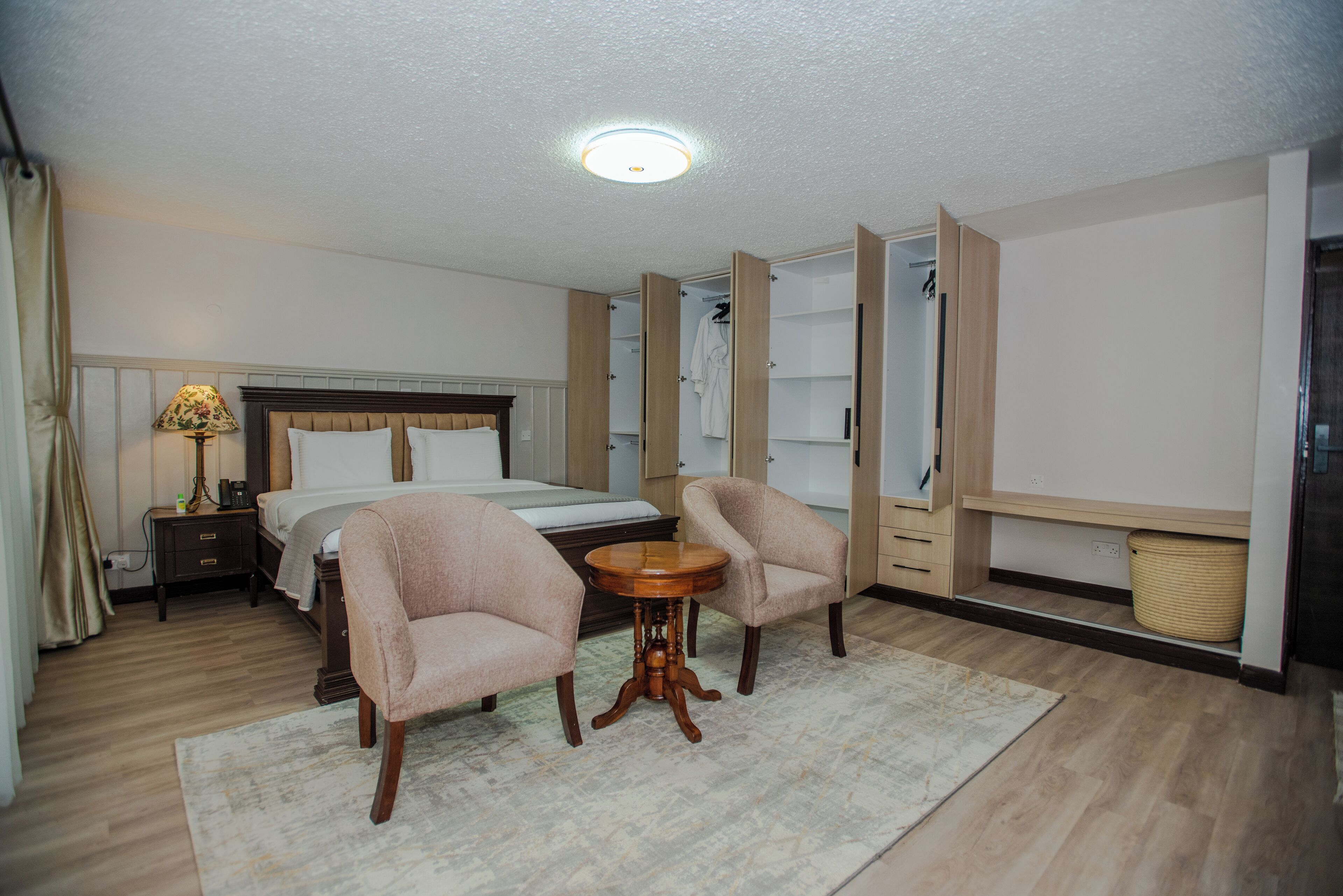 Une chambre d’hôtel comprenant un lit, deux fauteuils, une petite table ronde, une table de chevet avec une lampe et une armoire.