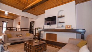 Een ruime woonkamer met een houten plafond, een open haard, een bank, een salontafel en een aan de muur bevestigde televisie.