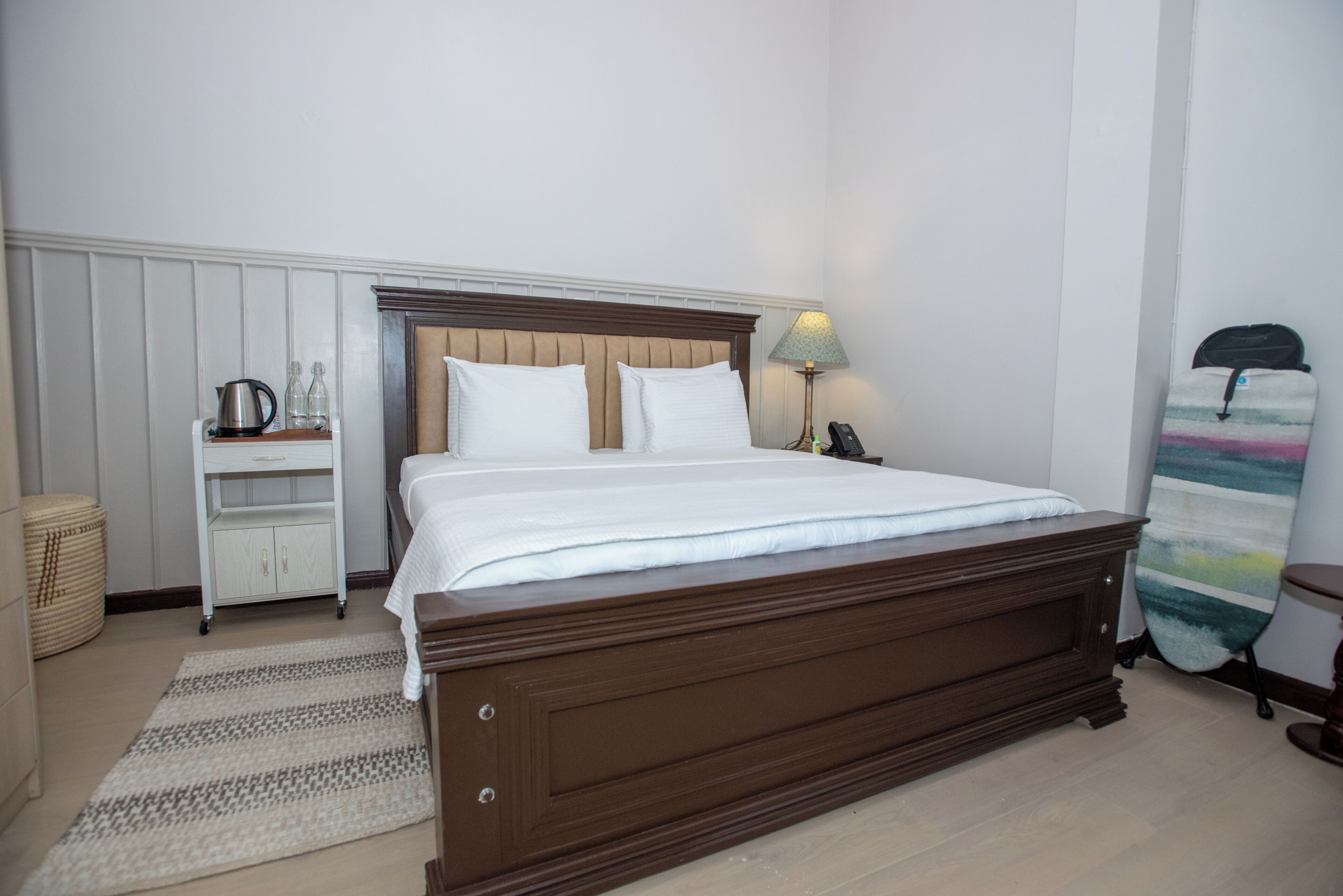 Una cama bien hecha con sábanas blancas, un cabecero de madera, una mesita de noche con una lámpara y un pequeño estante con una tetera y botellas.